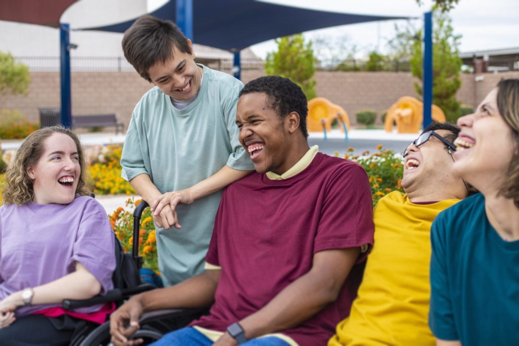 People laugh with their care worker in an outdoor community setting, sitting together and enjoying time in a garden space.