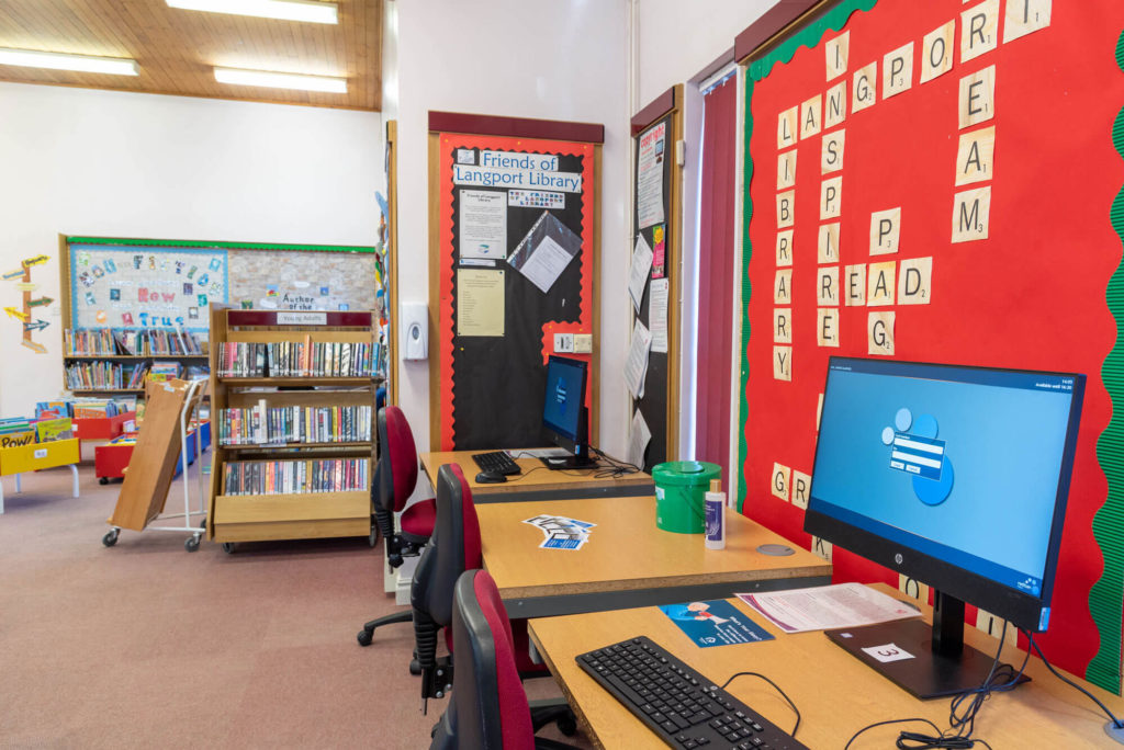 Public access computer area at Langport Library with desktop workstations, nearby book collections and a children's reading area in the background.