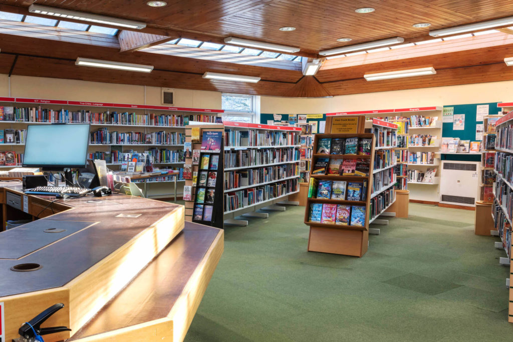 Main lending area at Wincanton Library with fiction and non-fiction book collections, featured book displays, customer service desk and wide aisles for browsing and borrowing.