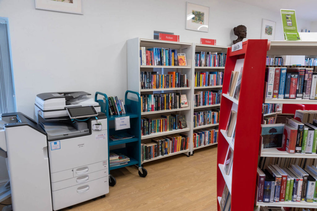 Printing and photocopying area at Street Library with a self-service multifunction printer, fiction book collections, display shelving and book trolleys for returned items.