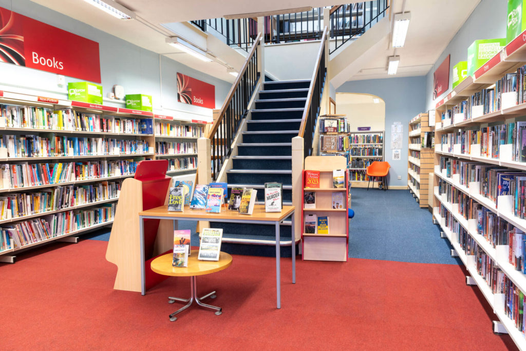 Books and fiction shelves at Cheddar Library, with featured book displays and a central staircase leading to the upper floor.