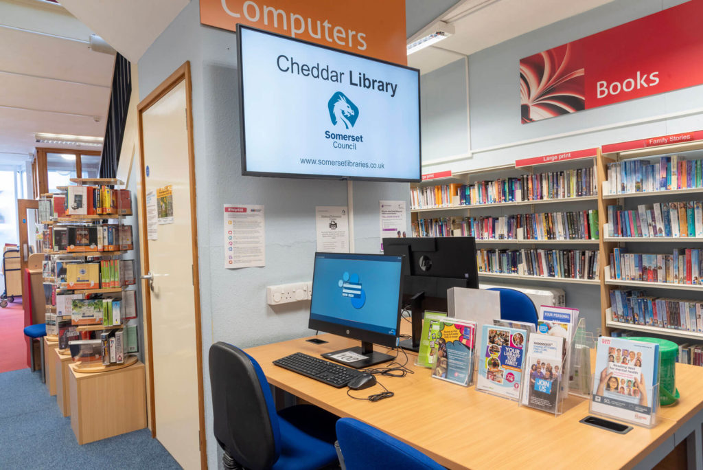 Public computer workstation at Cheddar Library with desktop PC, information leaflets and nearby bookshelves.