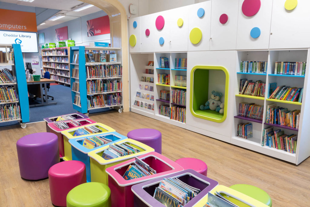 Children's library area at Cheddar Library with colourful book bins, low-level shelving, seating and a reading nook for young readers.