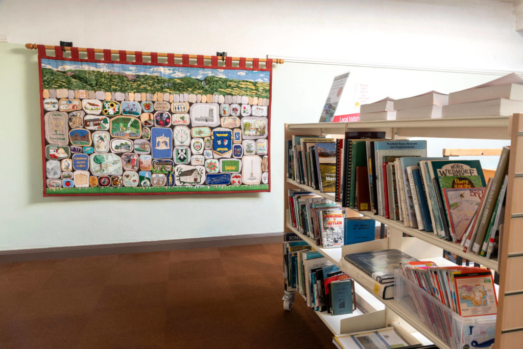 Local history section at Cheddar Library with shelves of reference books and a community artwork display on the wall.