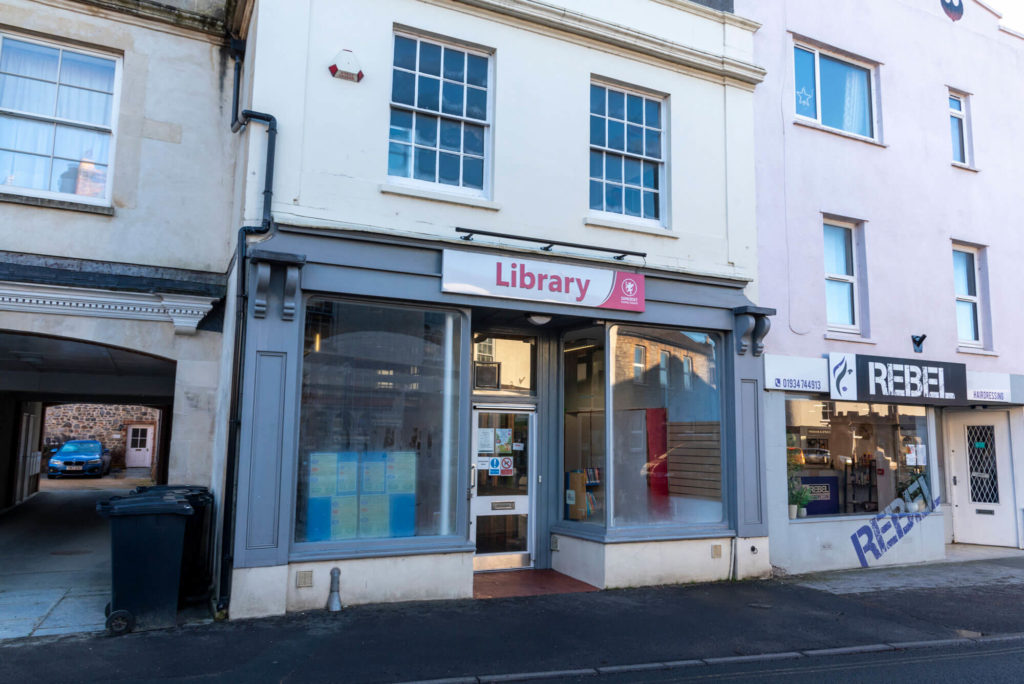 Exterior of Cheddar Library showing the main entrance on a high street, with large front windows and library signage above the door.