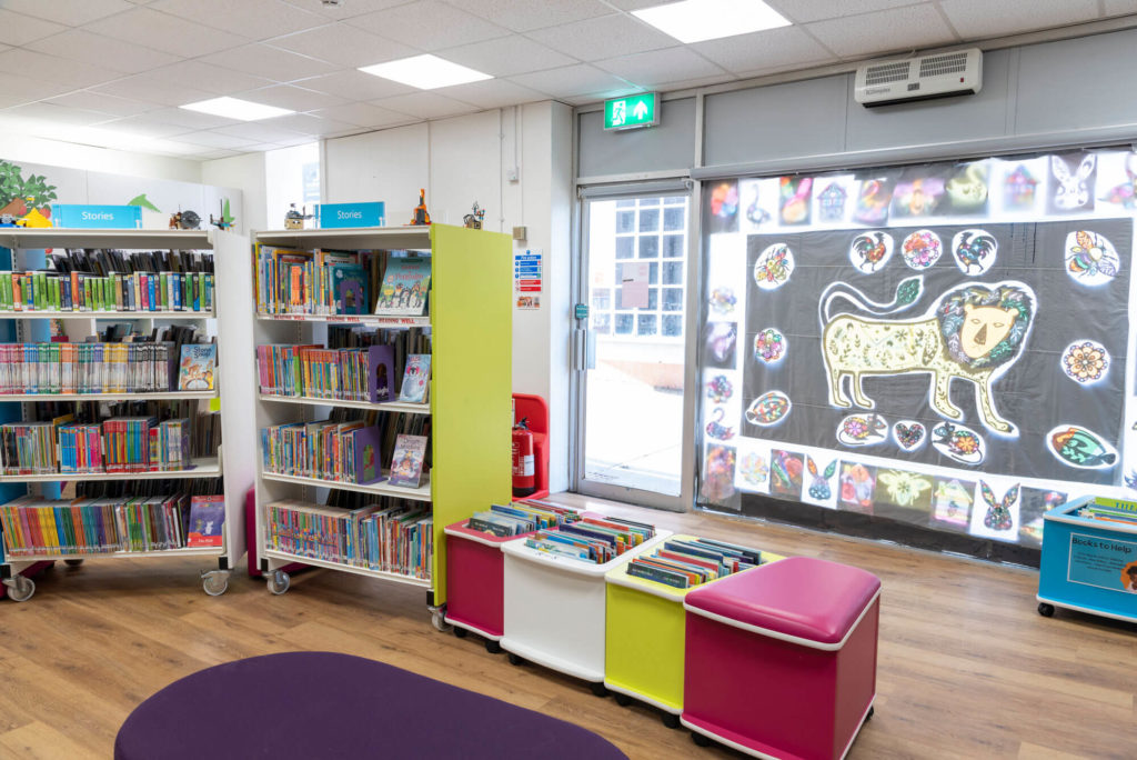 Children's area at Shepton Mallet Library with picture book collections, low-level book displays, colourful seating and a large window featuring children's artwork.