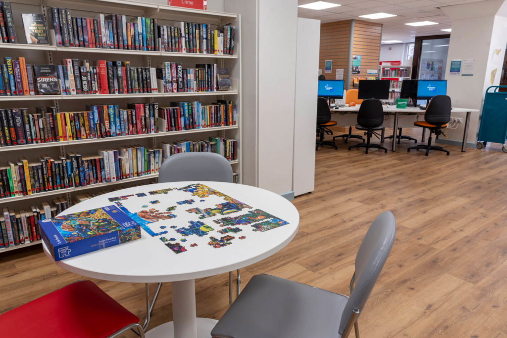 Community area at Shepton Mallet Library with a shared jigsaw puzzle table, crime fiction bookshelves and public computer workstations in the background.