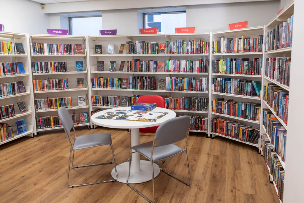 Community reading area at Shepton Mallet Library with a shared jigsaw puzzle table surrounded by fiction collections, including crime, fantasy and adventure books.