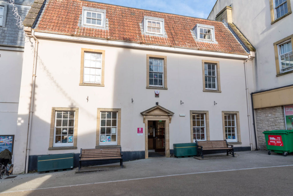 Exterior of Shepton Mallet Library, a historic two-storey building with a tiled roof, stone-framed entrance, benches and library signage in the town centre.