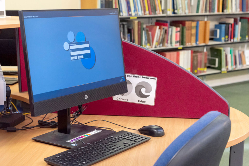 Public access computer workstation at Wells Library with desktop PC, keyboard, study desk and local history reference collections in the background.