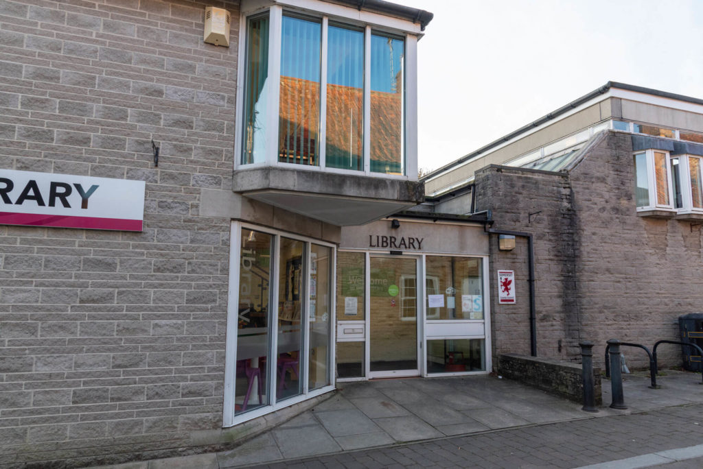 Exterior of Wells Library showing the main entrance within a modern stone building, with large windows, library signage and accessible pedestrian access from the town centre.
