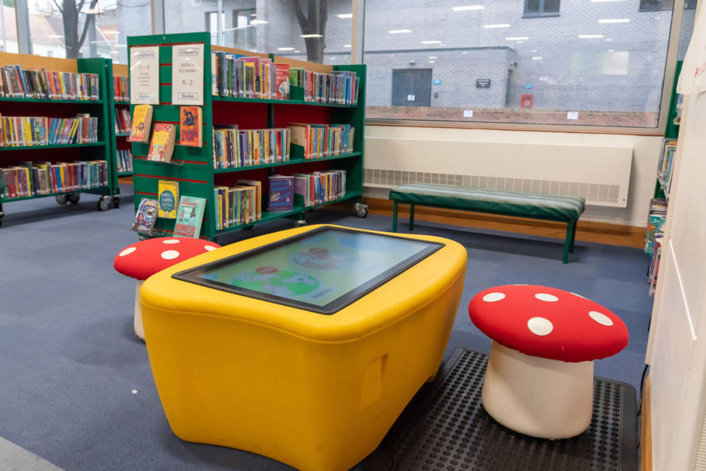 Interactive children's learning area at Taunton Library featuring a touchscreen activity table, mushroom-shaped stools, children's book collections and seating beside large windows.