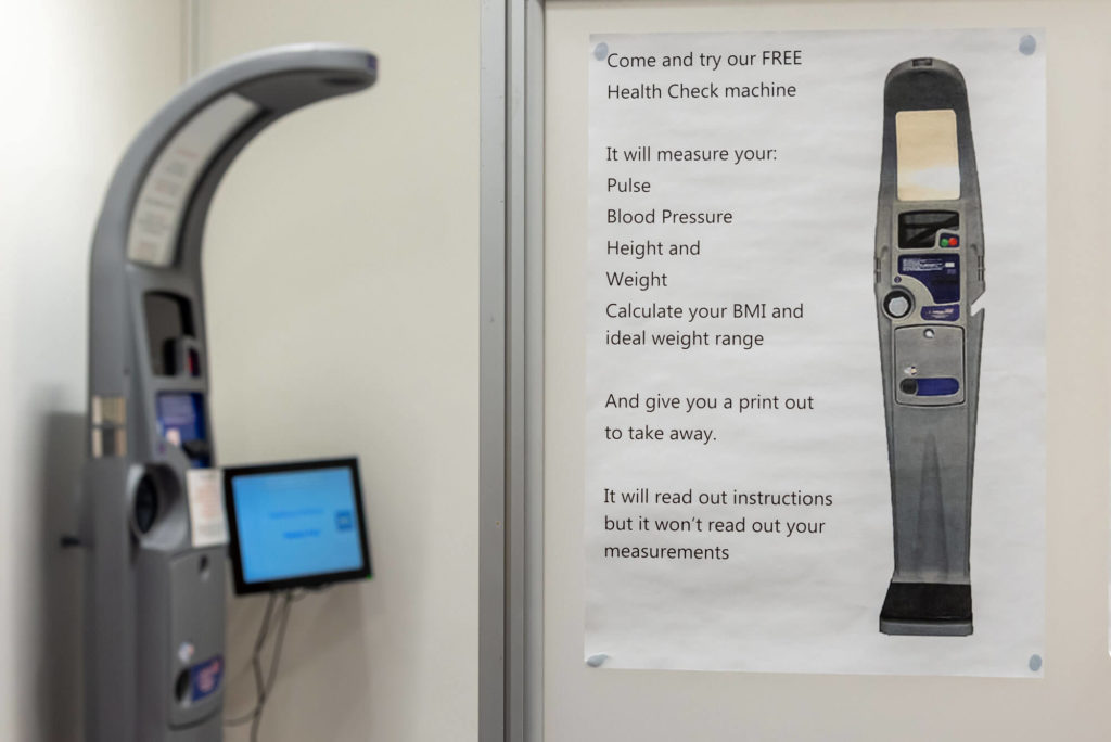 Free health check station at Taunton Library with equipment for measuring blood pressure, pulse, height and weight, alongside information explaining the health assessment service.