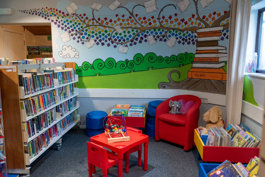 Children's reading area at North Petherton Library with picture book displays, colourful seating, activity table, toys and a large book-themed mural on the wall.