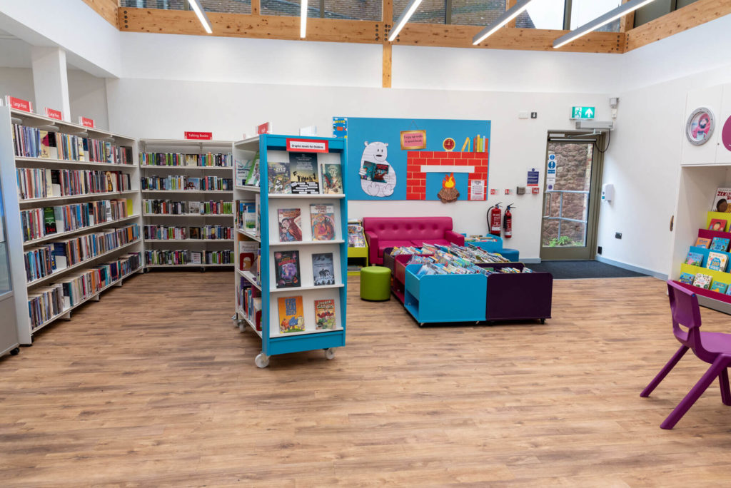 Open-plan children's and family area at Minehead Library with picture book displays, colourful seating, graphic novel collection and a bright modern interior with exposed timber beams.