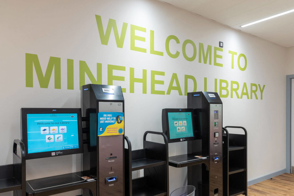 Self-service borrowing and returns kiosks at Minehead Library with touchscreen terminals and large welcome signage on the wall behind.