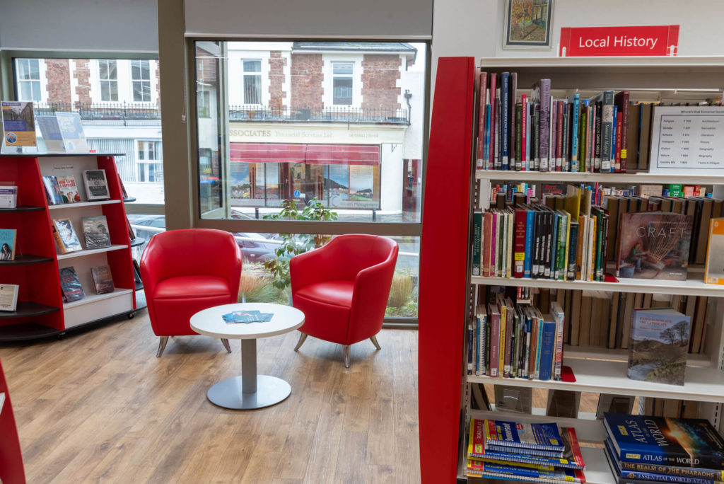 Local history area at Minehead Library with reference book collections, comfortable red seating, a small reading table and large windows overlooking the town centre.