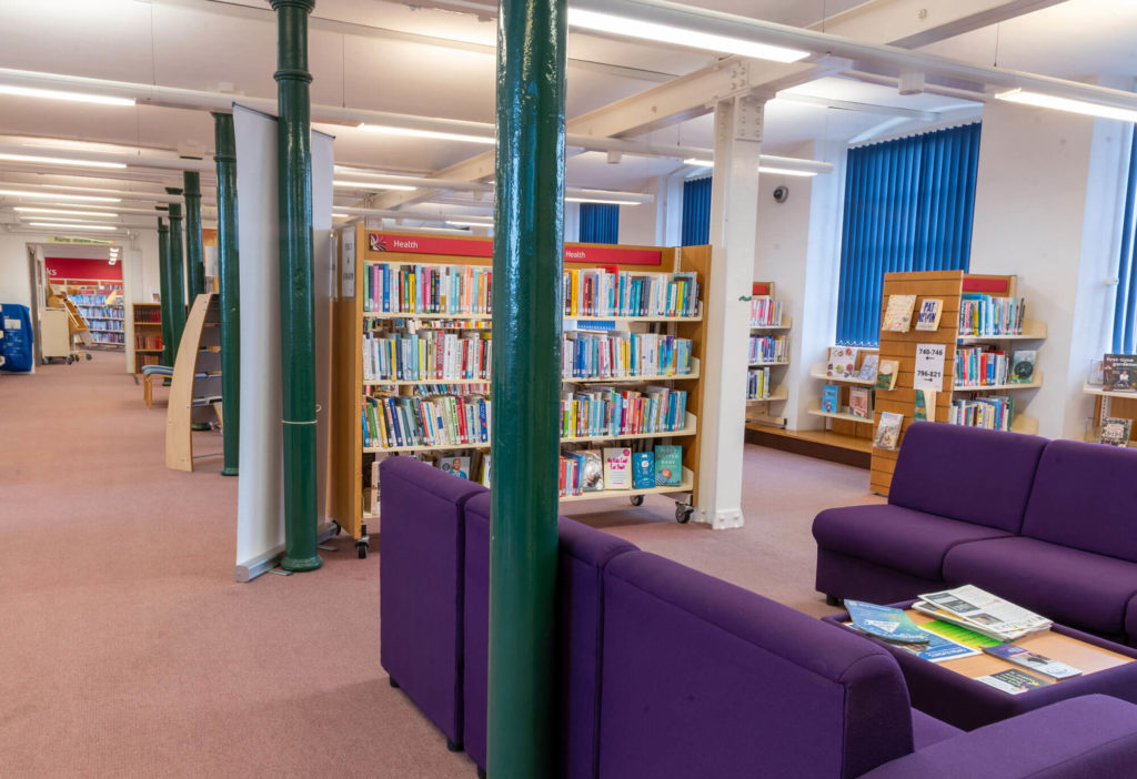 Library reading area with purple seating, health and wellbeing book shelves, and bright open study space.