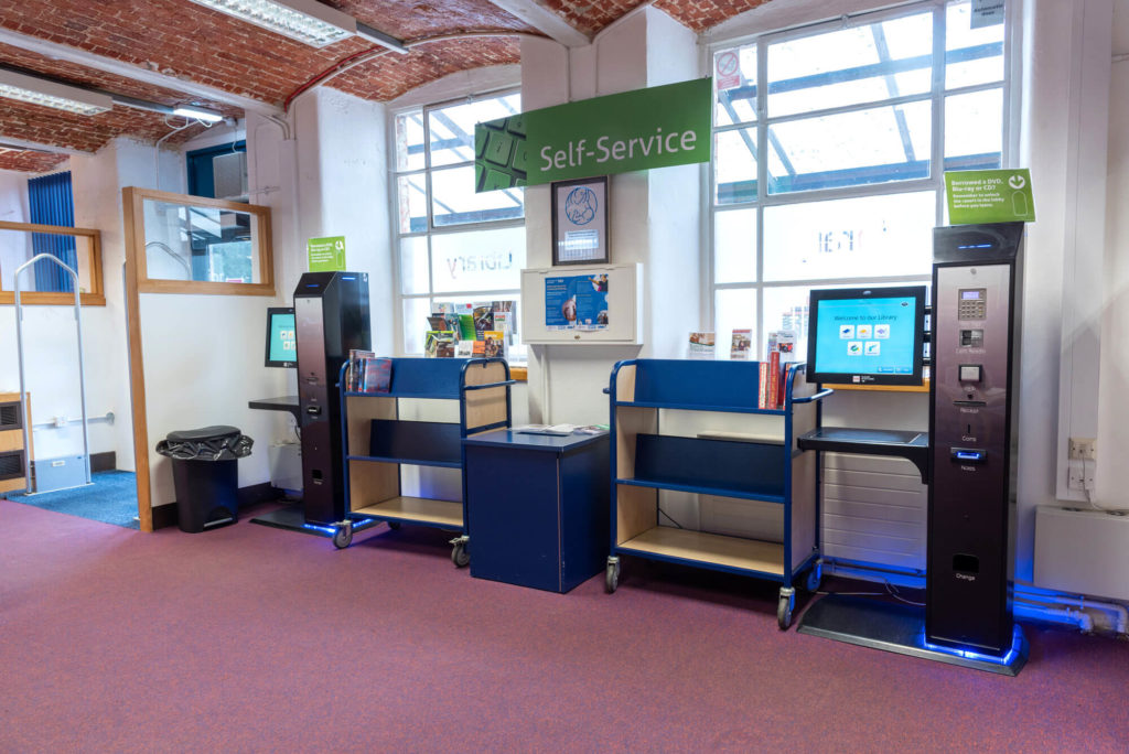 Self-service area at Chard Library with automated borrowing and returns machines, information displays and book trolleys.