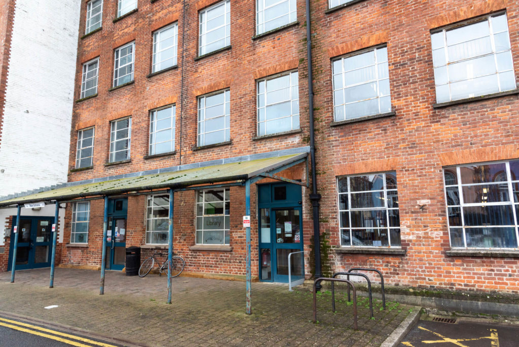 Exterior of Chard Library in a red brick building, showing the main entrance, covered walkway and bicycle parking.