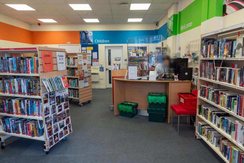 Main library area at Wiveliscombe Library with fiction and crime book collections, customer service desk, children's section and clearly labelled shelving for browsing and borrowing.