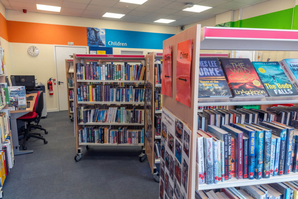 Fiction and crime book collections at Wiveliscombe Library with clearly labelled shelving, display books, public computer workstations and the entrance to the children's section in the background.