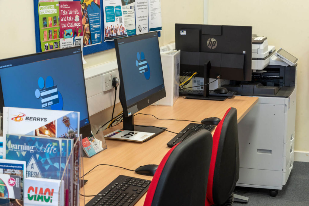 Public access computer area at Wiveliscombe Library with desktop workstations, seating, self-service printing and photocopying facilities, and information displays promoting local services and learning opportunities.