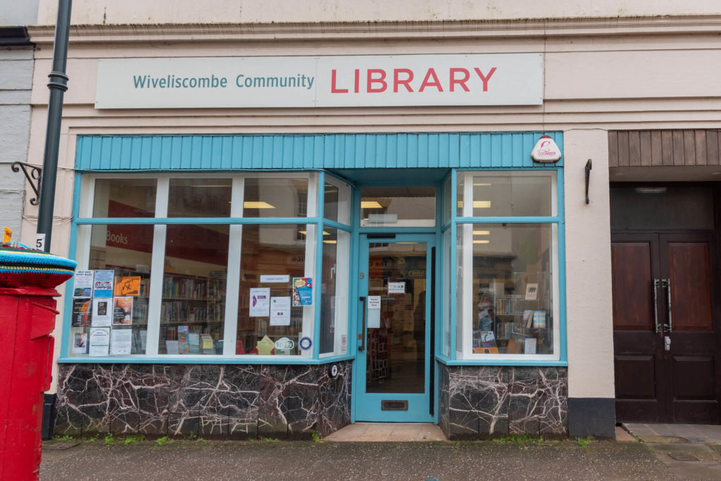 Exterior of Wiveliscombe Community Library showing the main entrance, large front windows, library signage and accessible access from the high street.