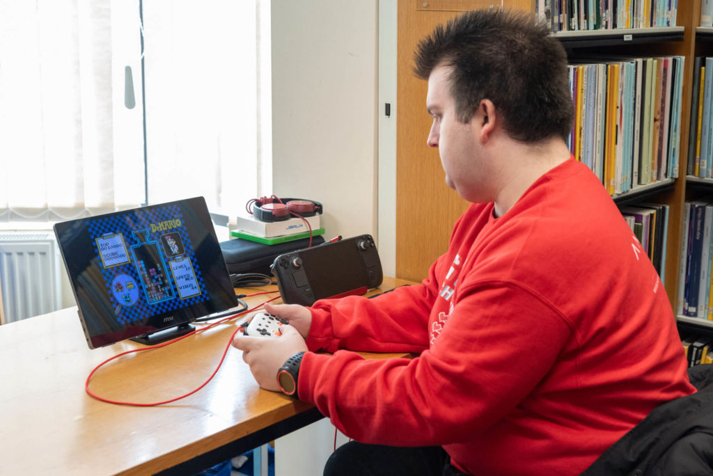 Accessible gaming and digital technology station at Yeovil Library with a tablet displaying a video game, handheld controller, headphones and shelving of books nearby.