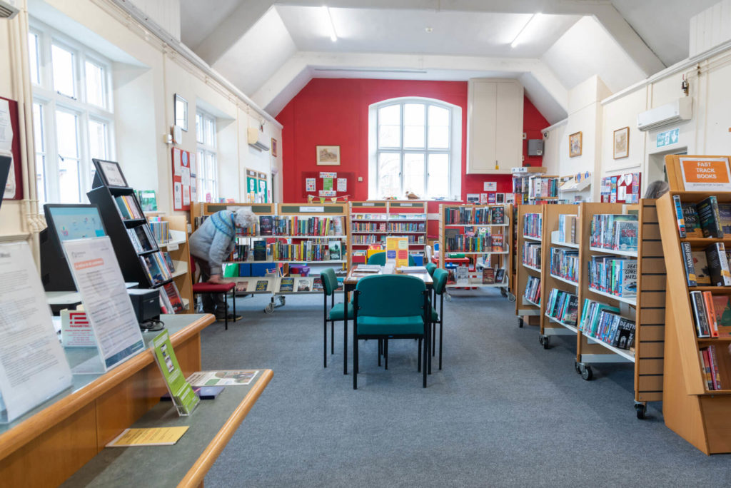 Main library area at Porlock Library with book collections, study chairs, browsing shelves, customer information displays and a bright reading space beneath a high vaulted ceiling.