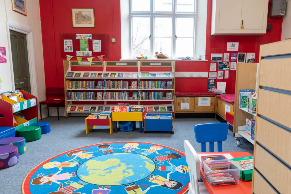 Children's area at Porlock Library with picture book collections, colourful display units, activity table, reading rug and seating for young readers and families.