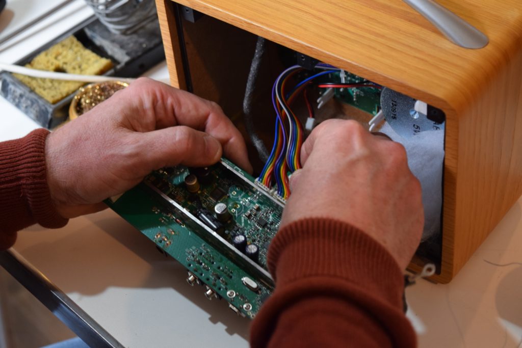A mans hands opening the front of a radio to expose the PCB and wires with a soldering iron in the background.