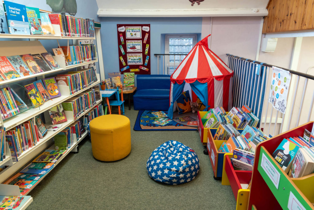 Children's reading area at Watchet Library with colourful picture book displays, bean bags, a circus-style reading tent, children's seating and shelves of books for young readers.