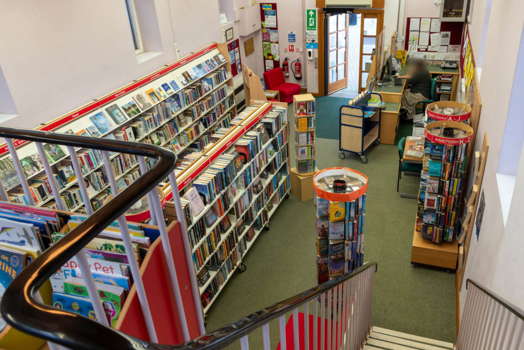 View across Watchet Library from the staircase, showing fiction and non-fiction bookshelves, browsing displays, customer service desk and the main entrance in a compact two-storey library.