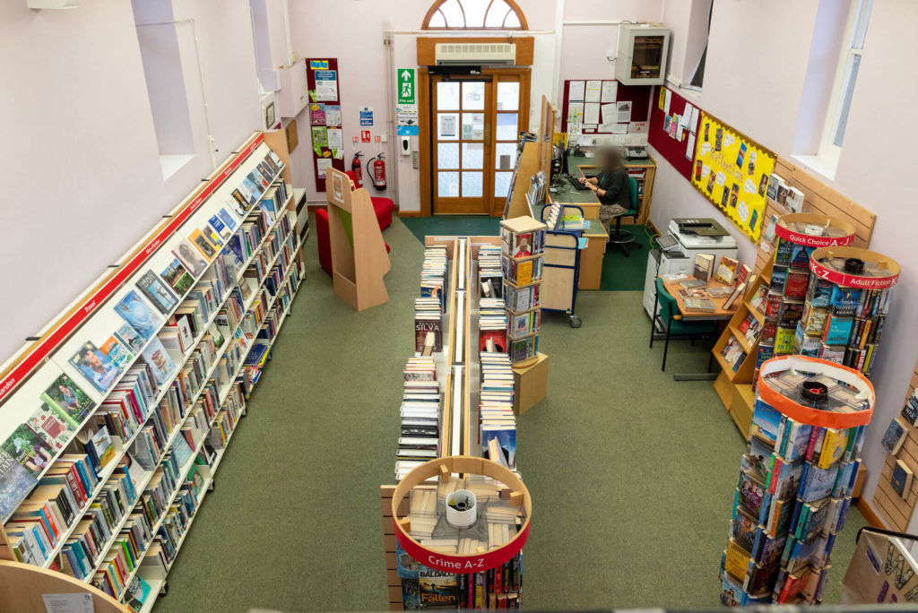 View across Watchet Library from the upper floor, showing fiction and non-fiction book collections, browsing shelves, customer service desk and the main entrance within a compact two-storey library.