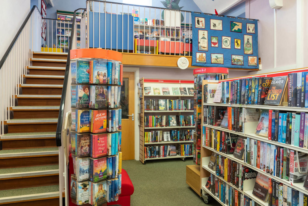 Fiction and crime collections at Watchet Library with browsing shelves, rotating book displays, staircase to the children's area and a textile artwork displayed on the upper gallery.