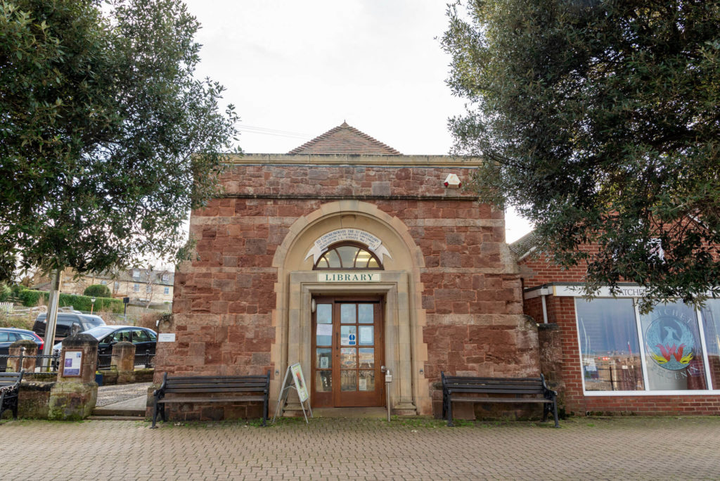 Exterior of Watchet Library, a historic red sandstone building with an arched entrance, library sign above the doorway and benches either side of the main entrance.