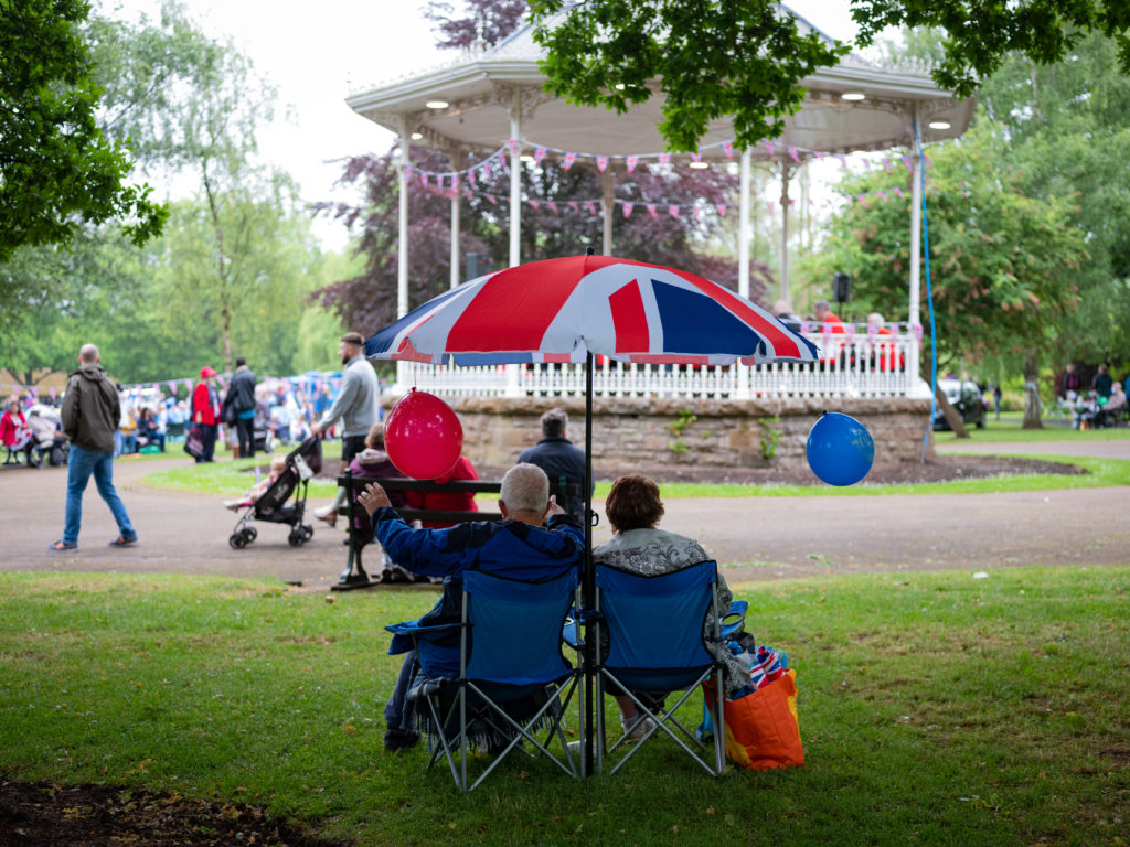 A couple sat on camping chairs under a union flag umbrella in Vivary Park Taunton , with the bandstand in the background. Taken at the Platinum Jubilee Celebration 2022.