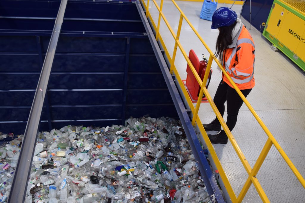 An employee in an orange hi-vis jacket and a blue hard hat stands on a balcony overlooking an area full of plastic that has been collected for recycling