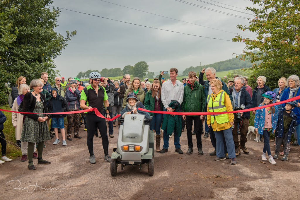 Group of people standing in front of red ribbon at the new path in Easton, while former HM Lord-Lieutenant of Somerset, Annie Maw cuts the ribbon.