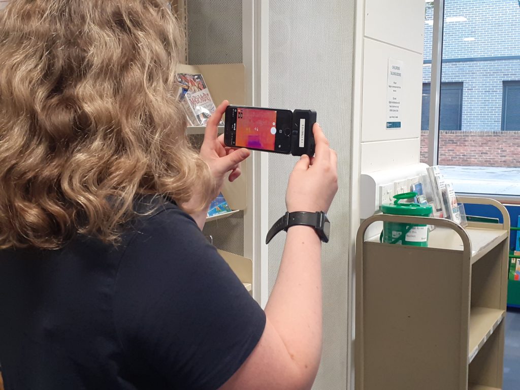 Person holding smartphone with a thermal imaging camera attached and the phone screen displaying the thermal image of the room.