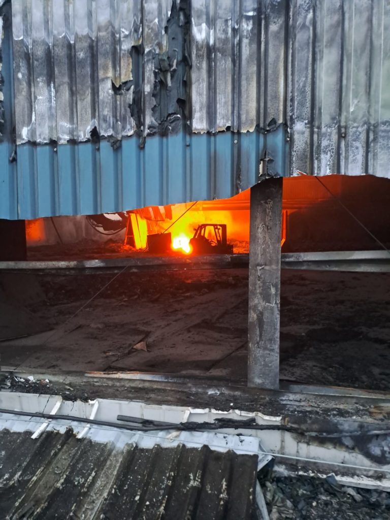 Looking through the side of the warehouse. Burnt and melted metal work reveals the burning fire in the background.
