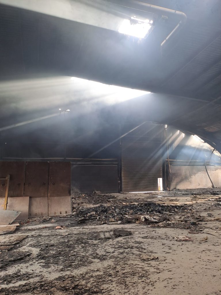 Light shines through the melted cladding on the material recovery facility. The area is fire damaged with ash over the ground.