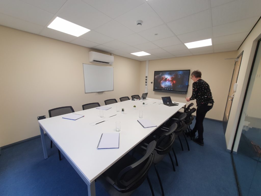 Bruton meeting room. There is a long desk with 10 chairs around it. There is a screen at one end mounted onto the wall, and a whiteboard on another wall.