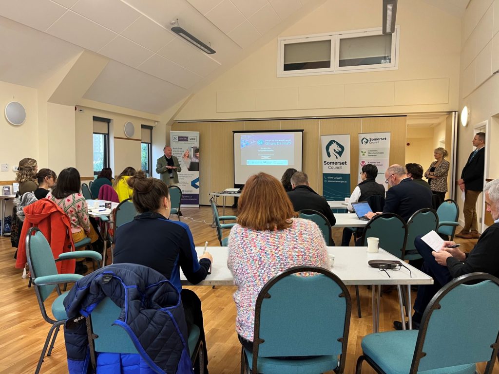 People sat around tables in a village hall listening to someone talking at the front.