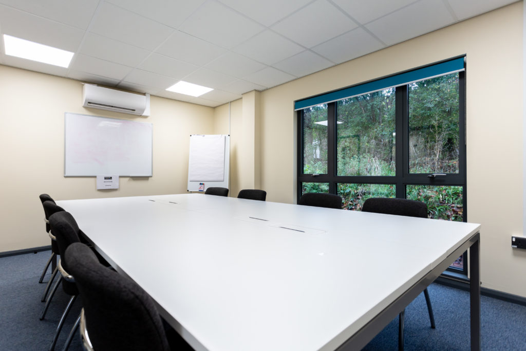 Wiveliscombe meeting space. There is a white desk with 8 chairs around it. There is a whiteboard on one wall, and a large floor to ceiling window on another.