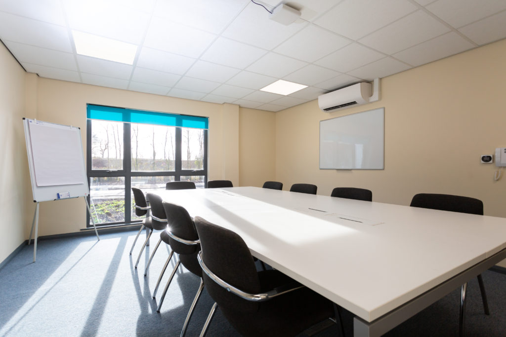 Wells meeting room. There is a large desk with 10 chairs around it. There is a floor standing flip chart white board in the corner next to a large floor to ceiling window. There is a whiteboard mounted on the wall.