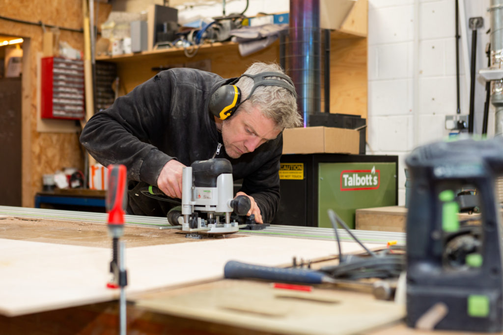 A man wearing ear protection is using router on a large sheet of wood clamped to a work bench