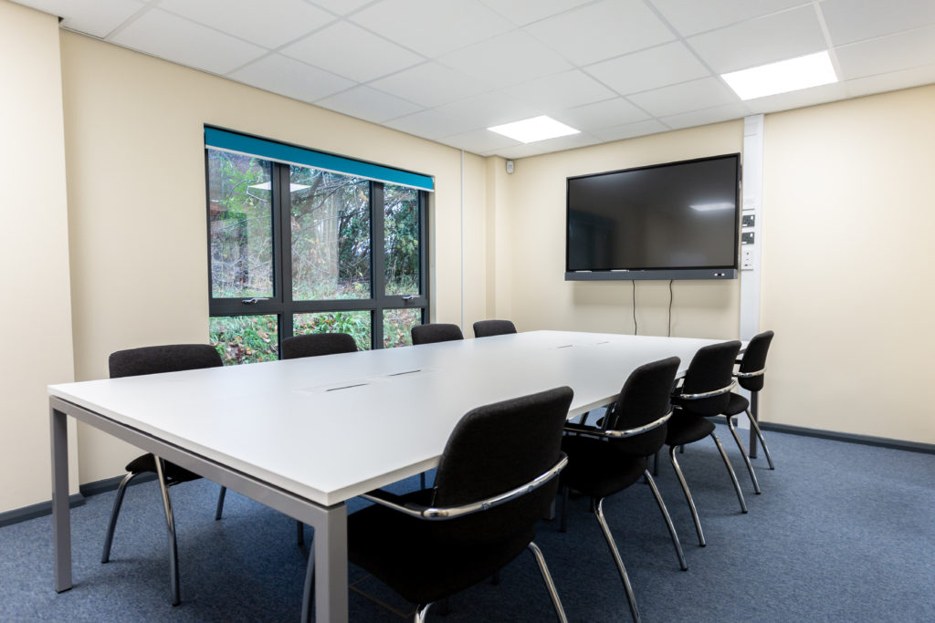 Wiveliscombe Meeting Room. There is a large desk with 8 chairs around it. There is a screen mounted on one wall. There are large floor to ceiling windows on another wall.