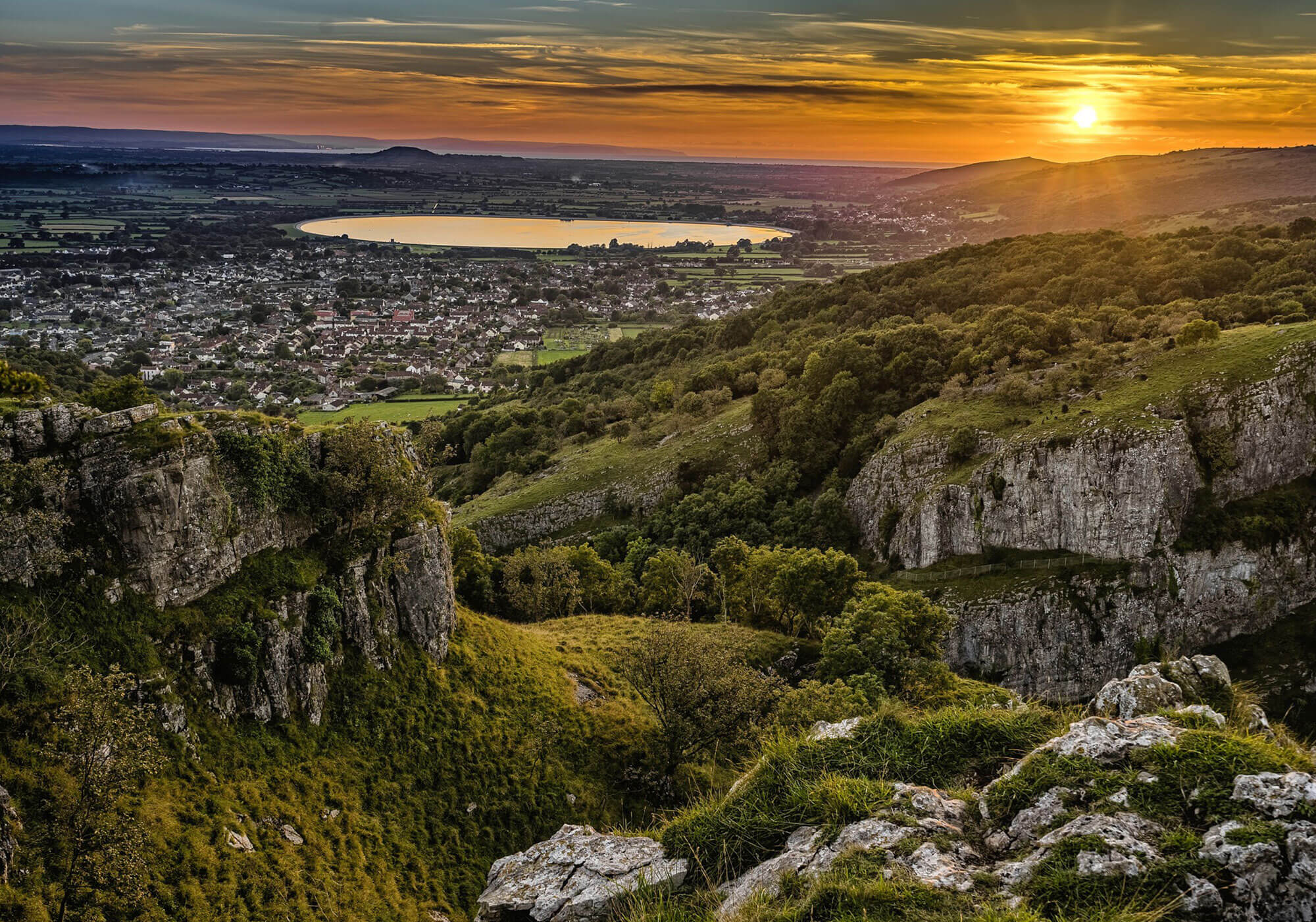 Photograph looking down at a Somerset town from high on the hills. The sun is setting in the distance.